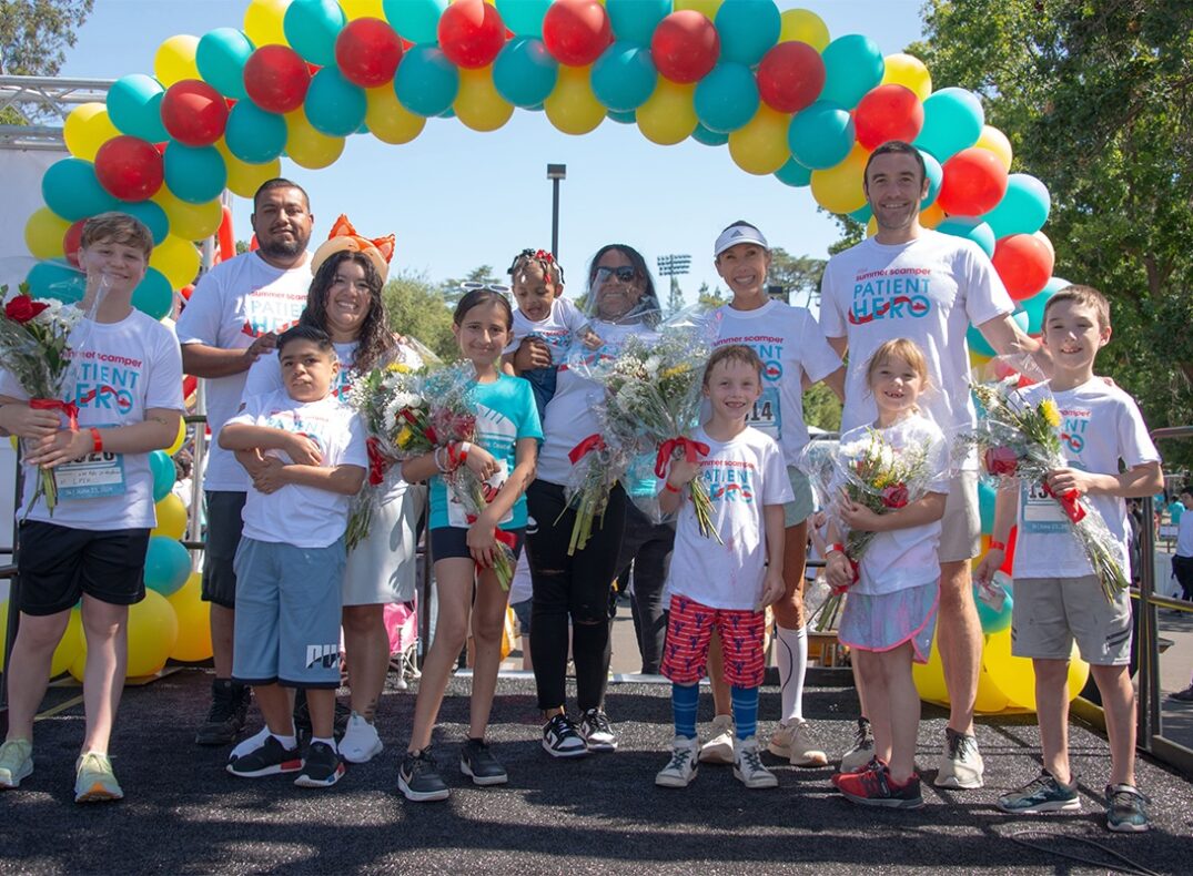 Patient hero families gather on stage under a balloon arch at Summer Scamper.