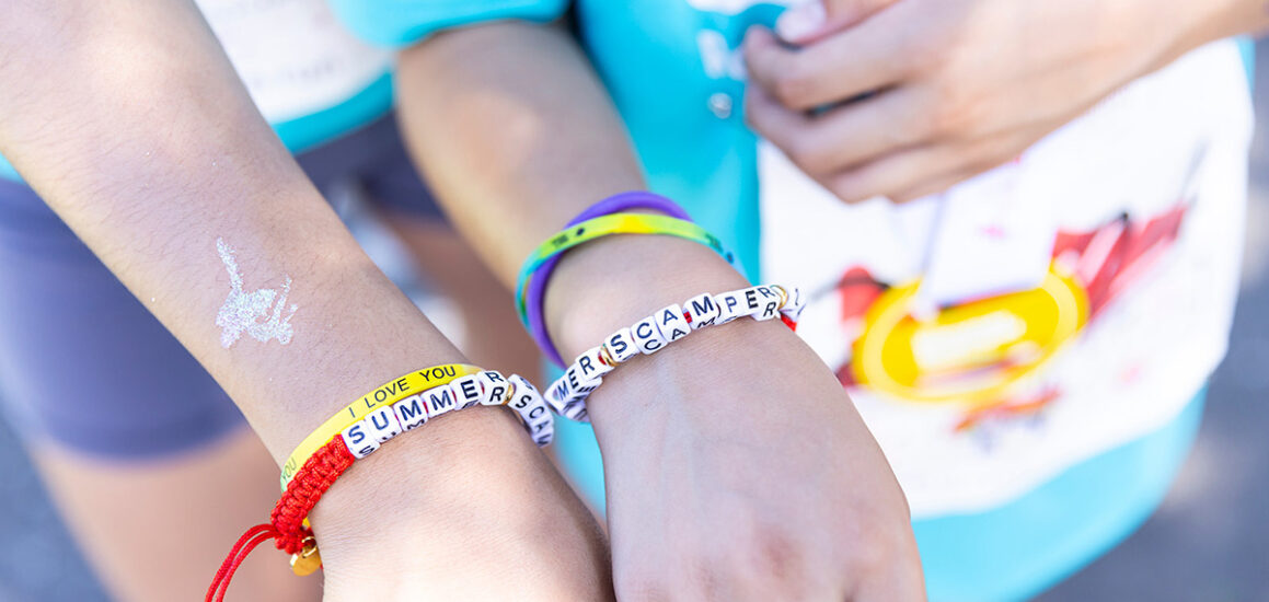 Two children's hands show off their homemade bracelets that say Summer Scamper.